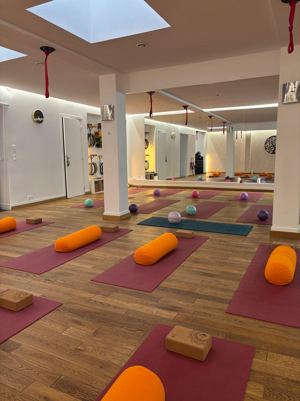 Salle lumineuse du studio sous verrière, tapis bordeaux, bolsters orange et ballons de Pilates